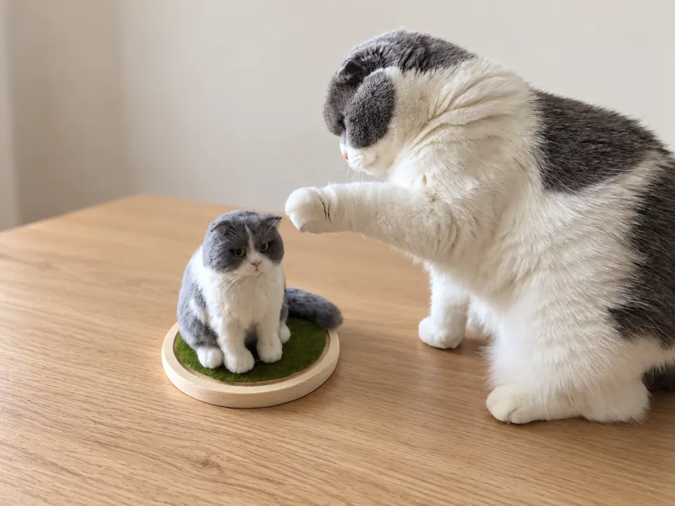 A grey-and-white cat reaching a paw towards a miniature needle-felted figure of itself standing on a round wooden base.
