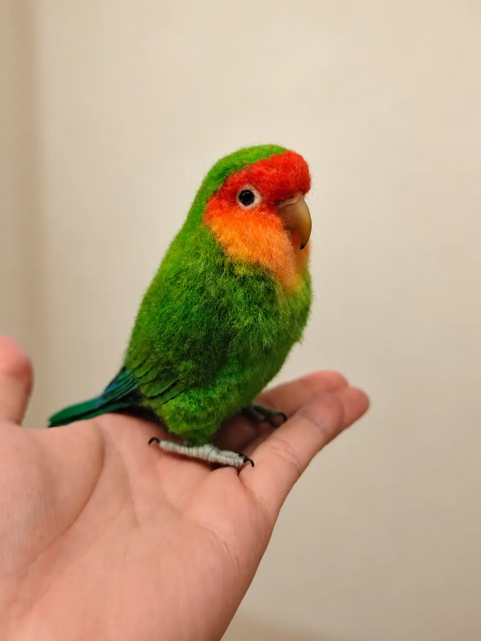 A needle-felted lovebird with a green body and an orange-red face, perched on an open palm.