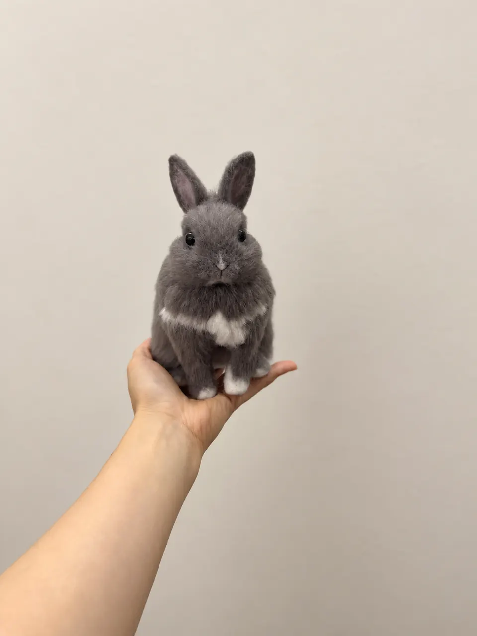 A needle-felted grey rabbit with upright ears and a white chest marking, sitting on an open palm.