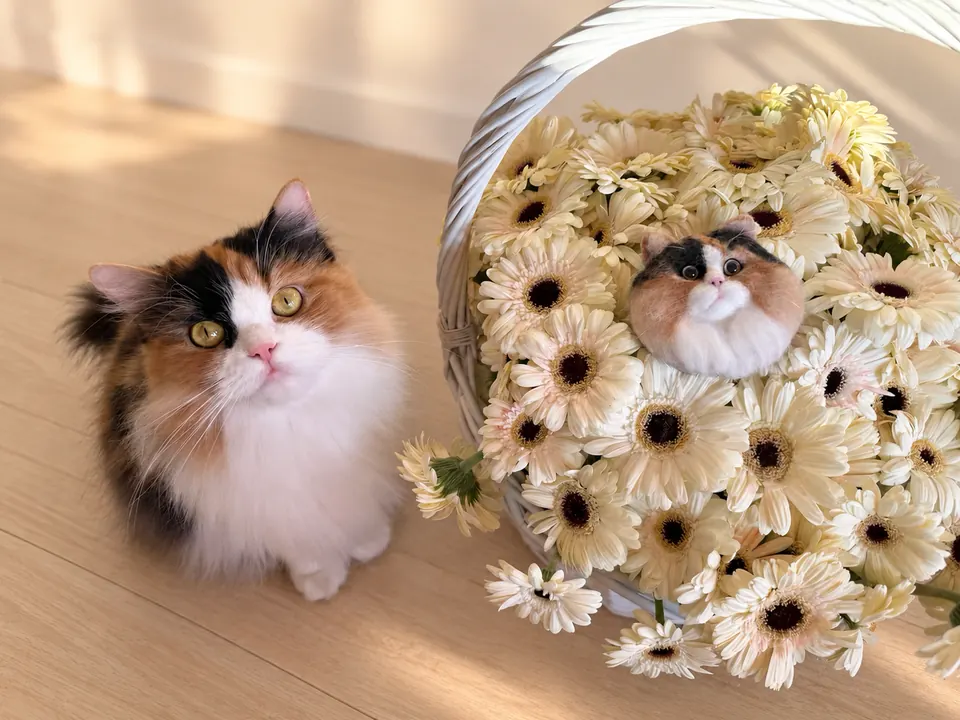 A calico cat sitting beside a white basket of daisies that holds a needle-felted portrait of a calico cat.