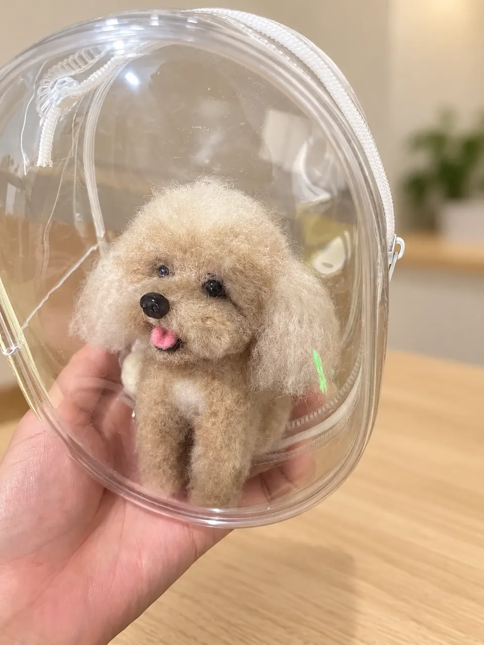 A cream needle-felted poodle with a pink tongue inside a clear zipped display case, held in one hand.