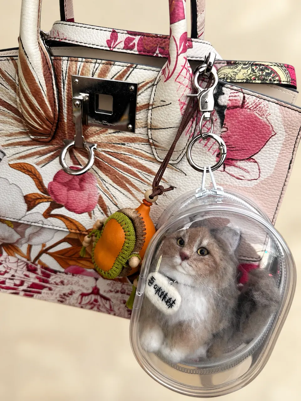 A needle-felted cat inside a clear oval case, clipped to the handle of a patterned handbag as a charm.