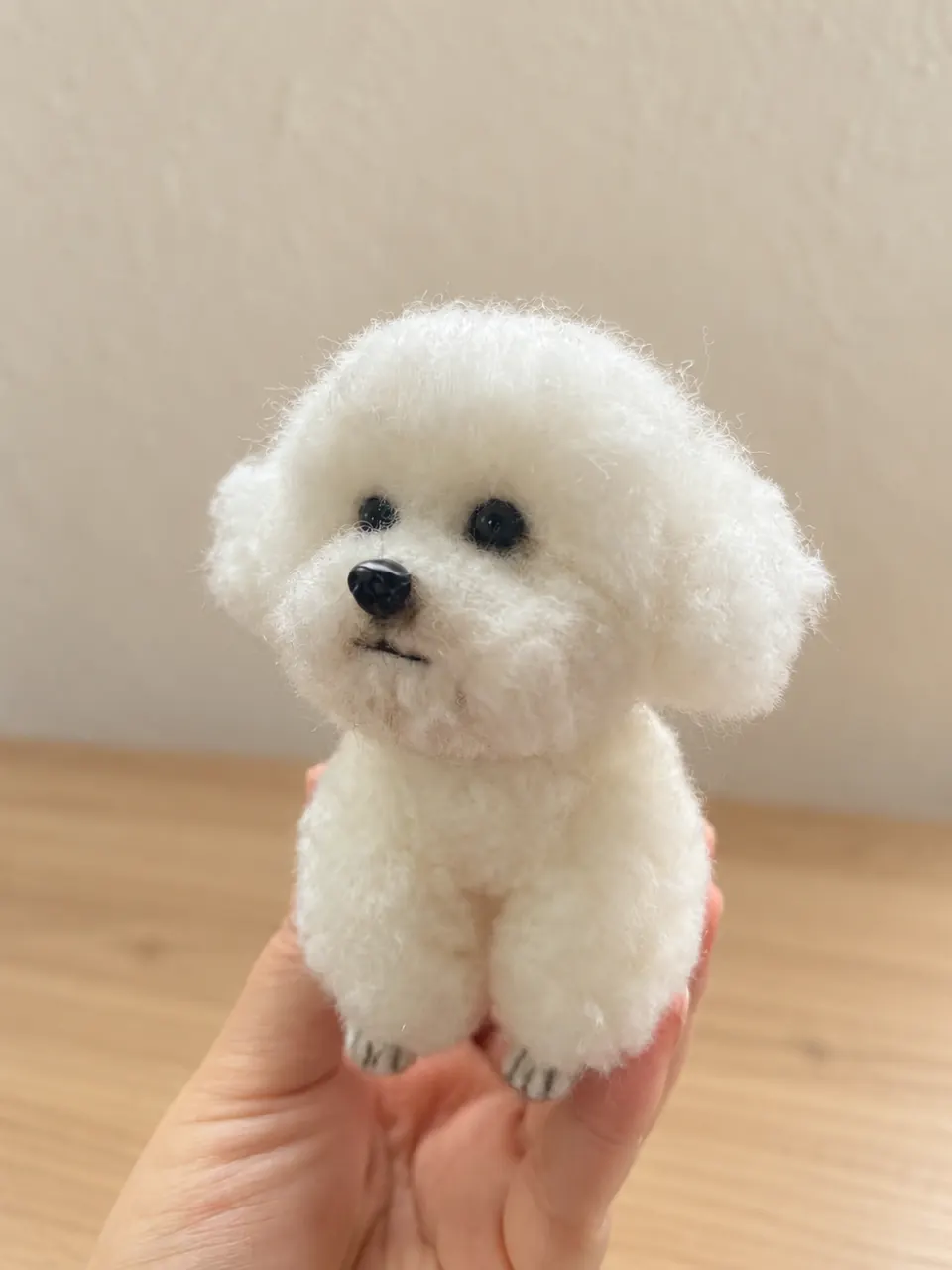 A needle-felted white bichon-type dog standing in a cupped hand, with a fluffy rounded coat and a dark nose.